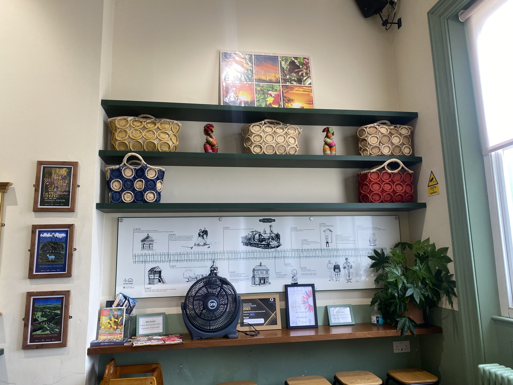 Inside Café at the Station: sage-green shelves of handwoven baskets above the Bat and Ball heritage timeline, framed vintage railway posters on the wall