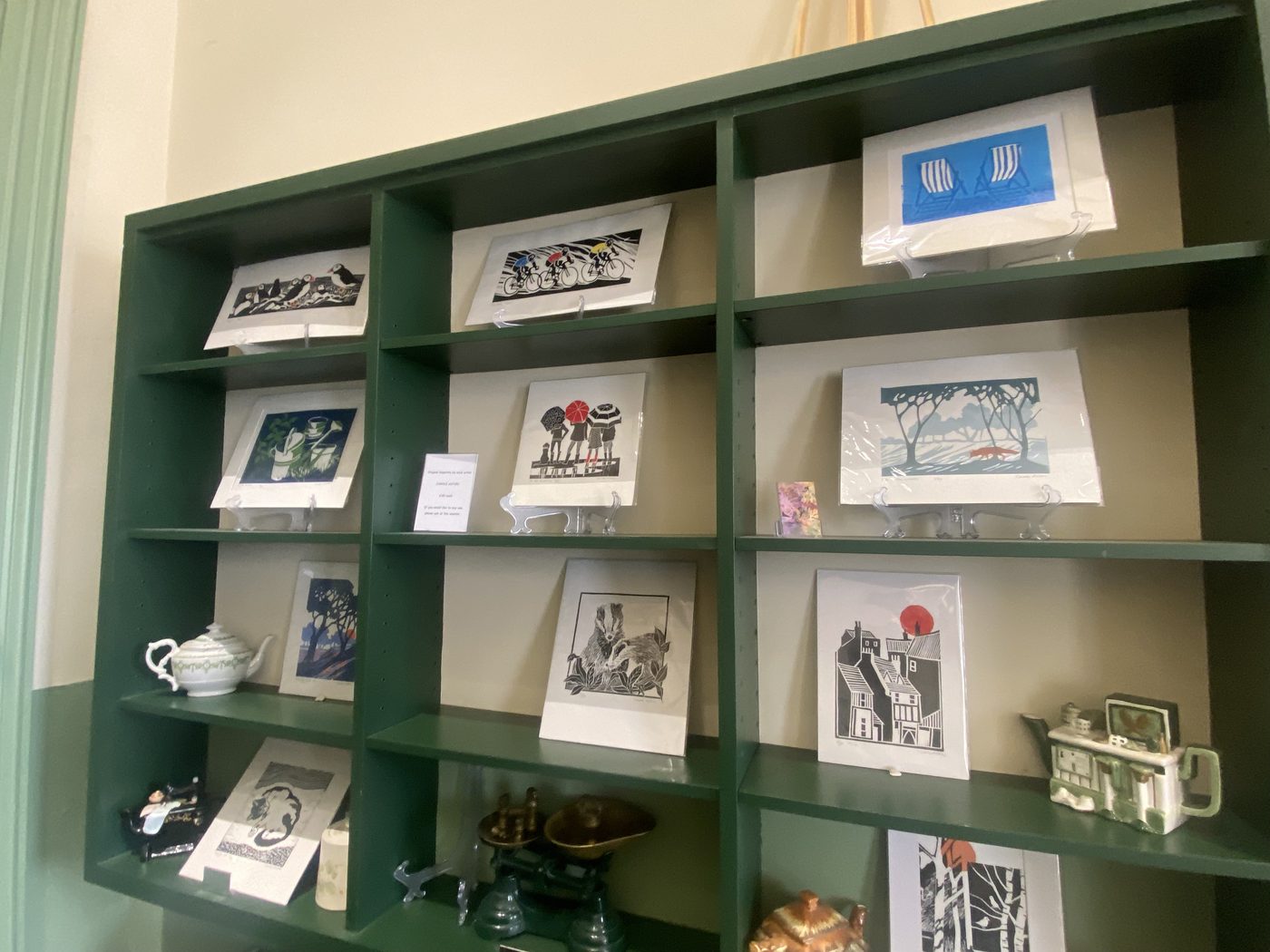 More linocut prints on green cubby shelves with brass scales and a ceramic cottage teapot