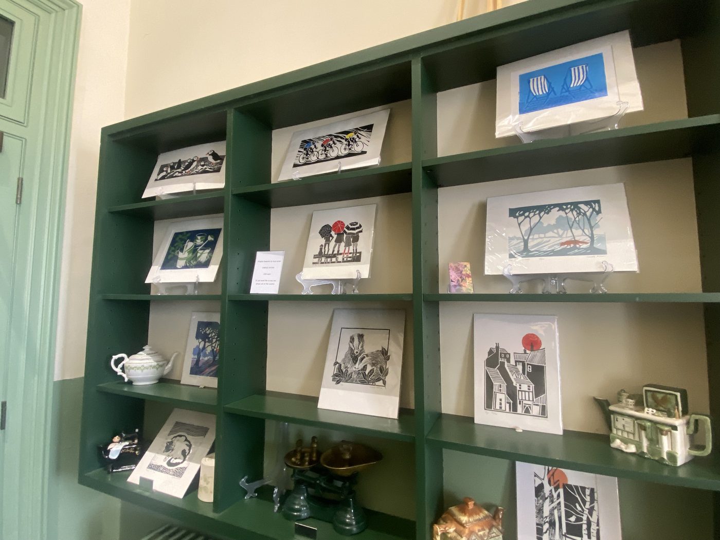 Hand-printed linocut art cards on a sage-green shelf: puffins, cyclists, blue deckchairs, a woodland badger and houses under a red sun
