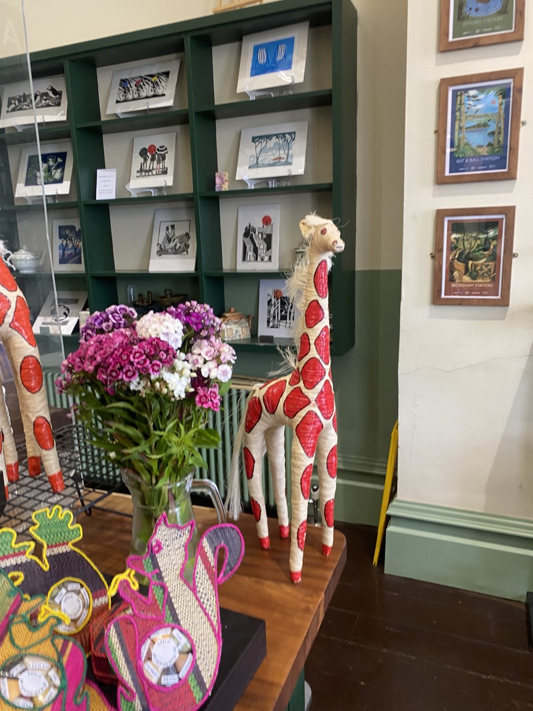 A handwoven giraffe figure and colourful woven animals beside fresh flowers, with linocut cards and vintage railway posters behind